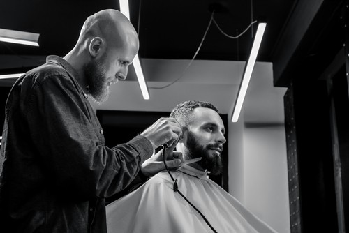 ALD BARBER IS TRIMMING THE HAIRCUT OF HIS BEARDED SERIOUS CLIENT. HE IS USING A HAIR CLIPPER